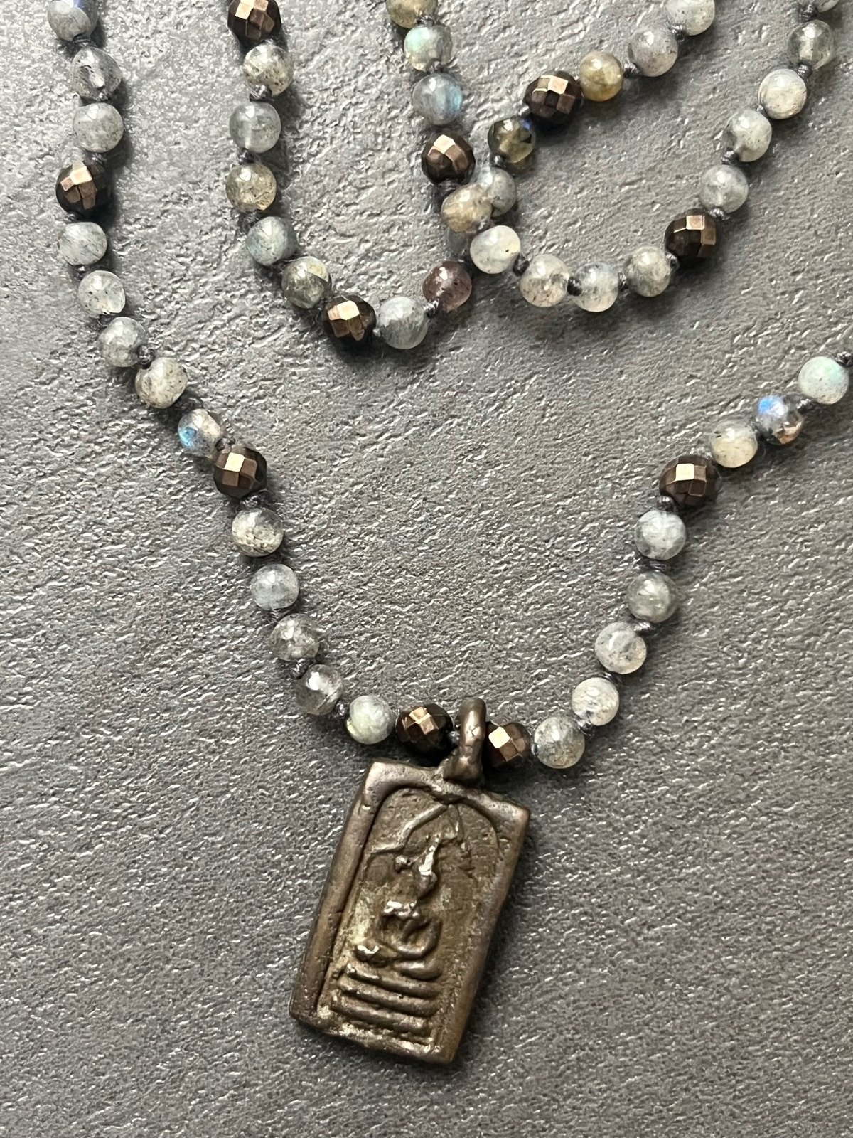 buddha spark necklace featuring a double-faced thai buddhist amulet in antique bronze finish with faceted bronze-colored hematite beads hand-knotted with round labradorite beads, as a perfect blend of style and spirituality