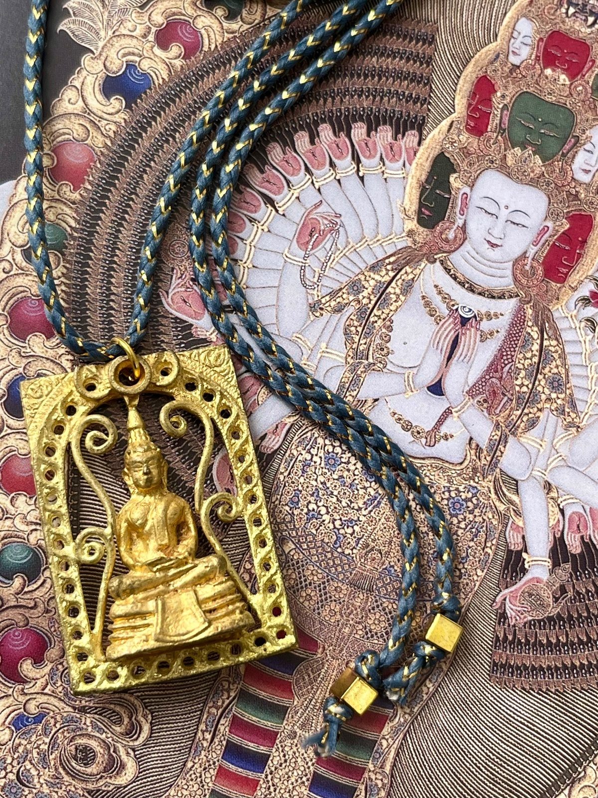 necklace with grey blue golden lurex cotton cord and thai golden brass amulet depicting a seated buddha with ornate frame, symbolizing protection, peace, and spiritual guidance